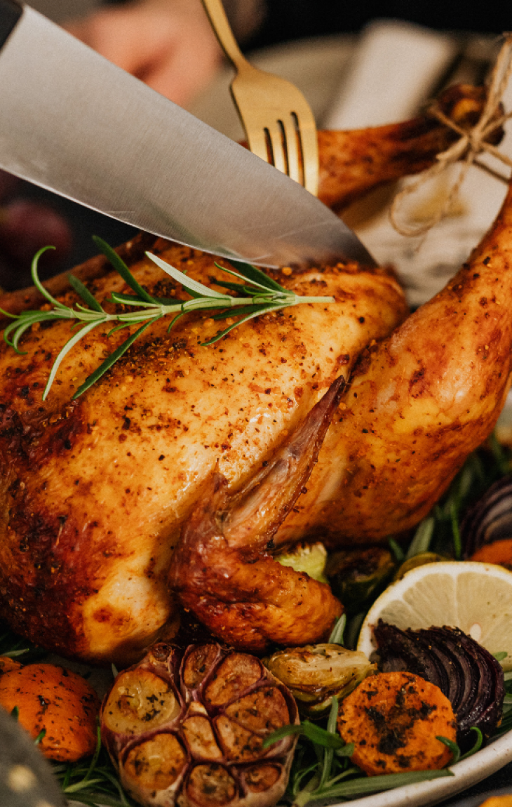A person carving a roasted chicken garnished with herbs, lemon slices, and vegetables on a dinner table.