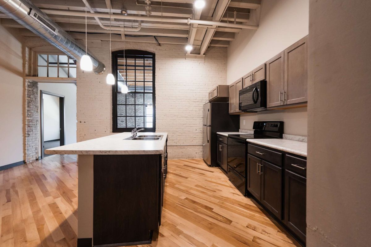 Modern kitchen with dark cabinets, stainless steel appliances, island, wood floors, and exposed ceiling pipes.