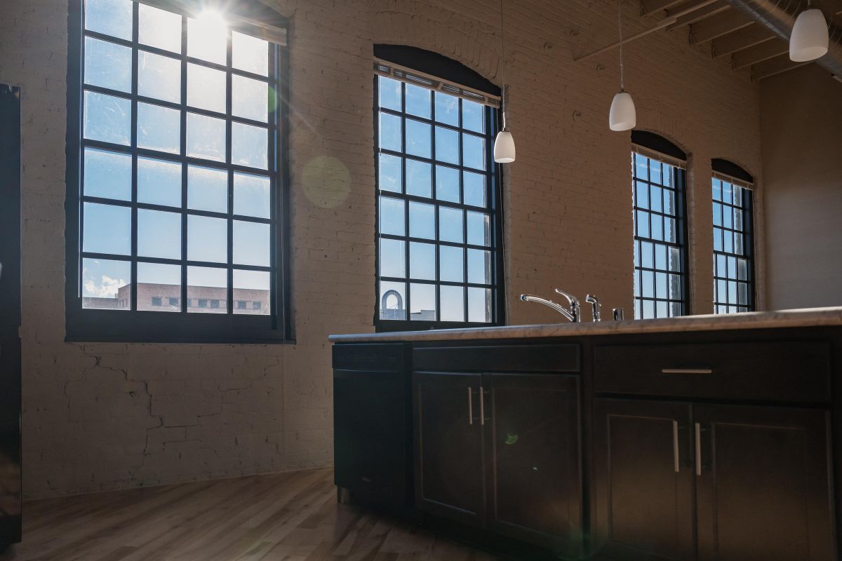Sunlight streams through large industrial windows in a modern kitchen with dark cabinets and pendant lights.