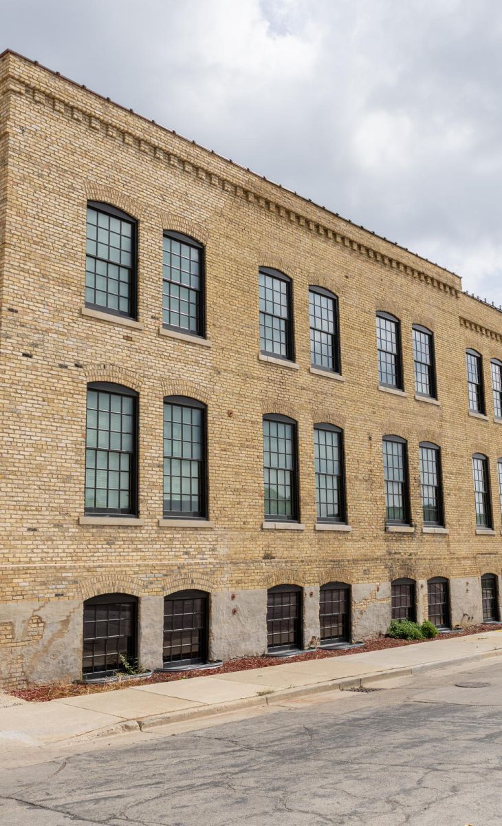 A large, tan brick industrial building with many black-framed windows on a quiet, empty street.