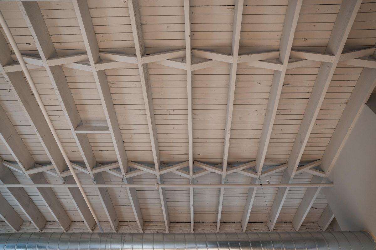 Exposed wooden ceiling beams with white paint and a metal air duct running along the bottom.