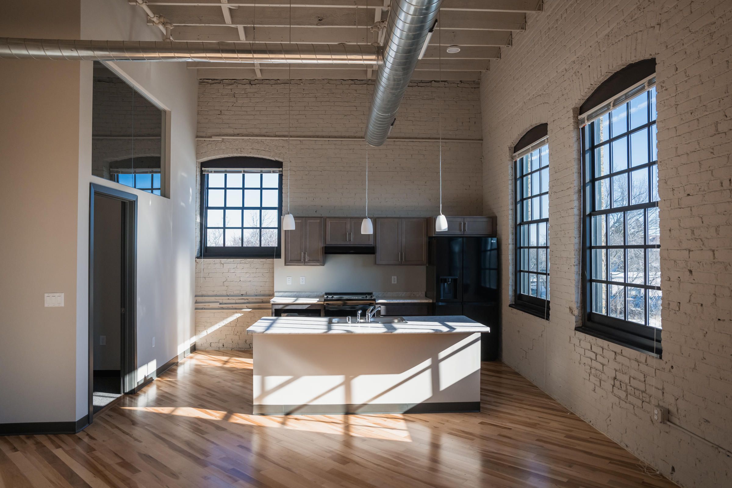 Modern loft kitchen with large windows, exposed brick walls, wood floors, and industrial-style ceiling.