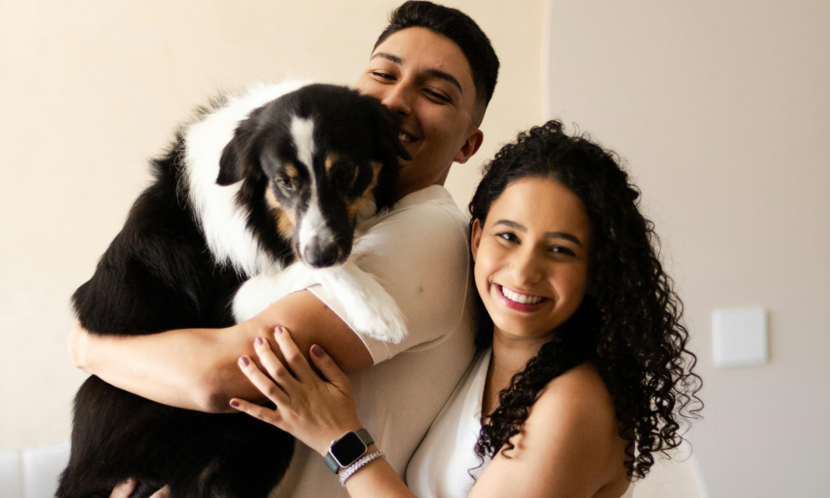 Smiling couple poses together, man hugging a black and white dog, woman stands beside them, all looking happy.