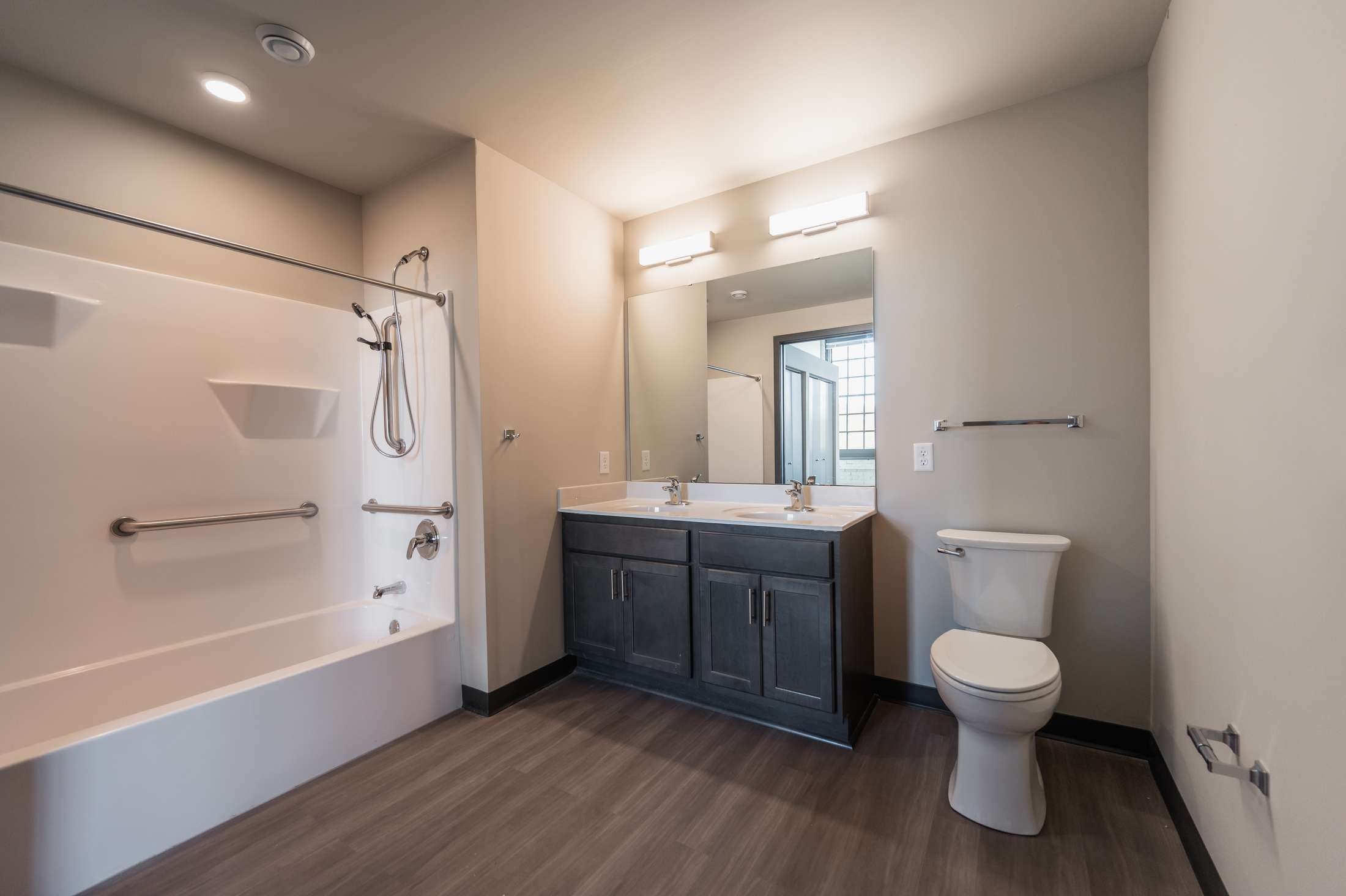 Modern bathroom with a bathtub, grab bars, large mirror, double sink vanity, and toilet.