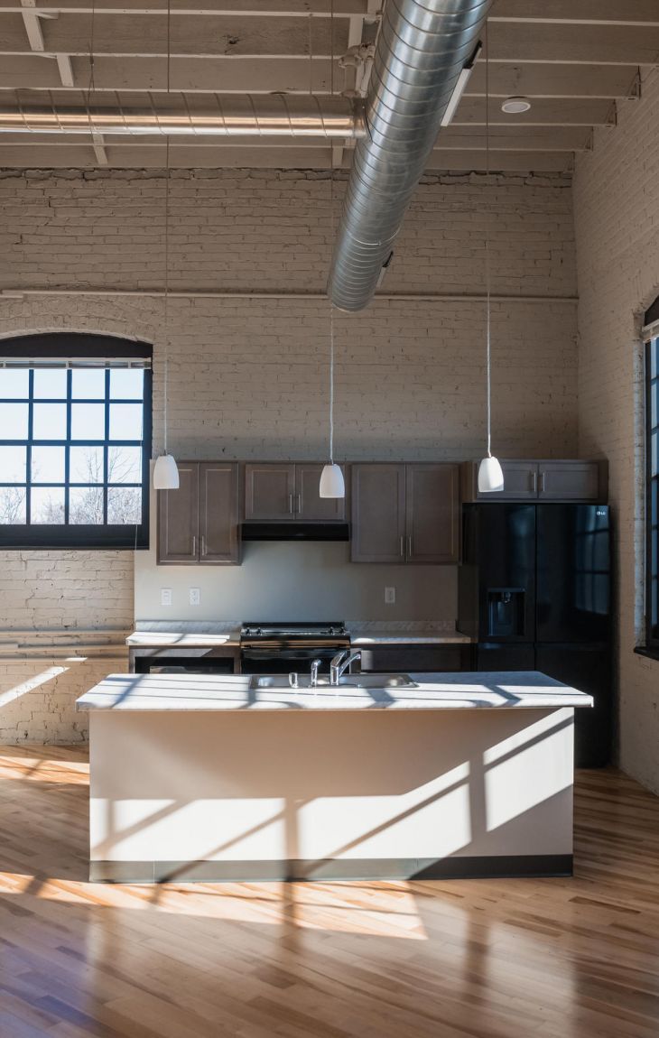 Modern loft kitchen with large windows, exposed brick walls, wood floors, and industrial-style ceiling.