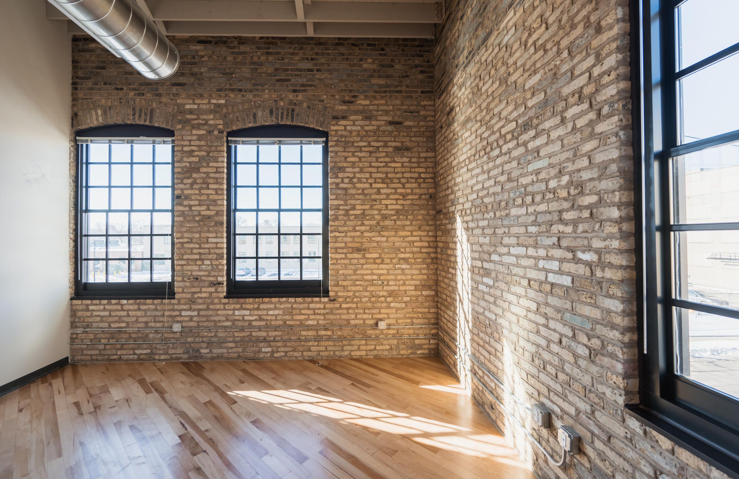 Sunlit loft room with brick walls, large black-framed windows, and light hardwood floors.