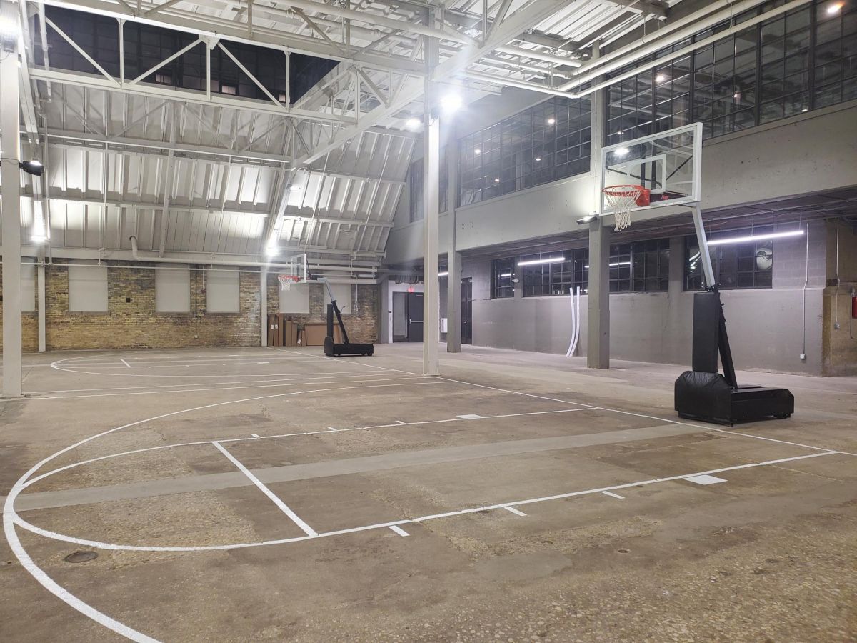 Indoor basketball court with two hoops, marked lines, high ceilings, and exposed brick and metal walls.