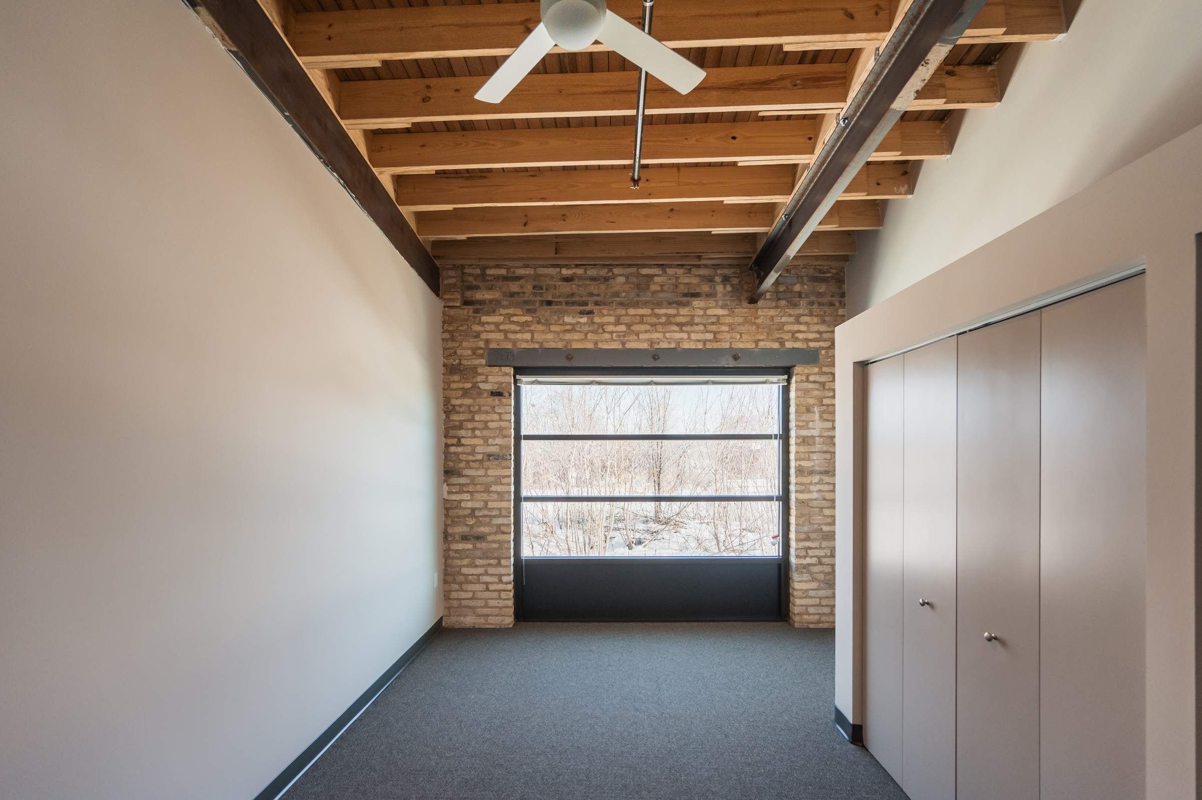 A modern empty room with exposed wooden beams, a brick wall, large window, and closed double-door closet.