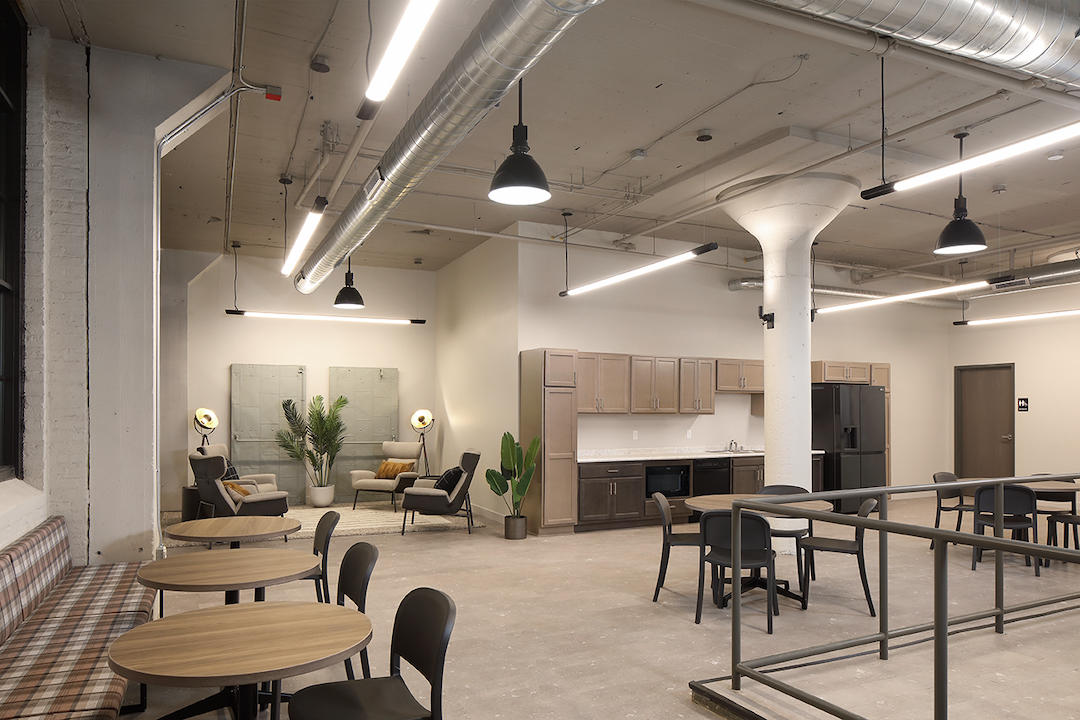 Modern office lounge with tables, chairs, kitchenette, plants, and industrial-style lighting and ceiling.