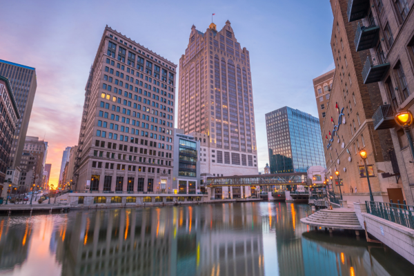 Downtown city buildings reflected in a river at sunset, with a pedestrian bridge over the water.