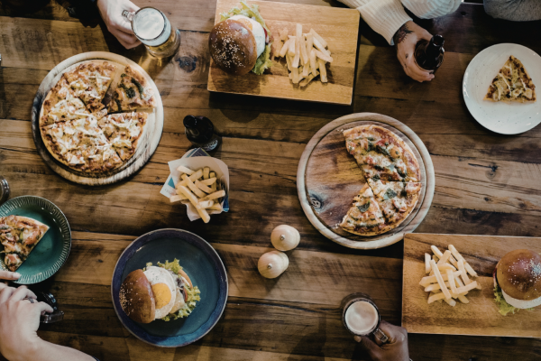 A table with pizzas, burgers, fries, and drinks, with people’s hands reaching for food.